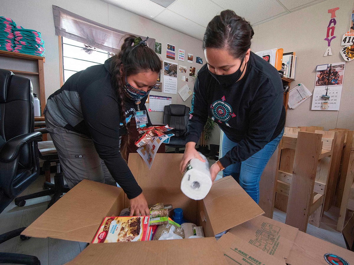 Two women in black hoodies and masks on place a paper towel roll and assorted food items into a cardboard box with small window in background February 2022.