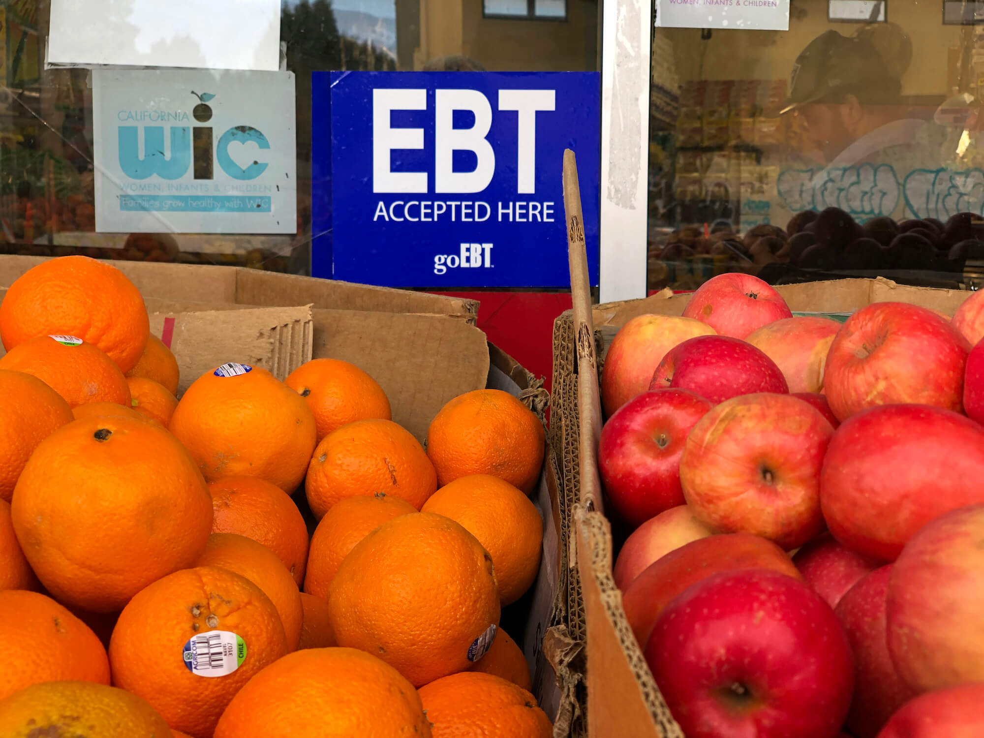 Orange and apples in cardboard boxes outside of a grocery store window. A blue EBT accepted here sticker next to a California WIC sticker in the window. December 2019